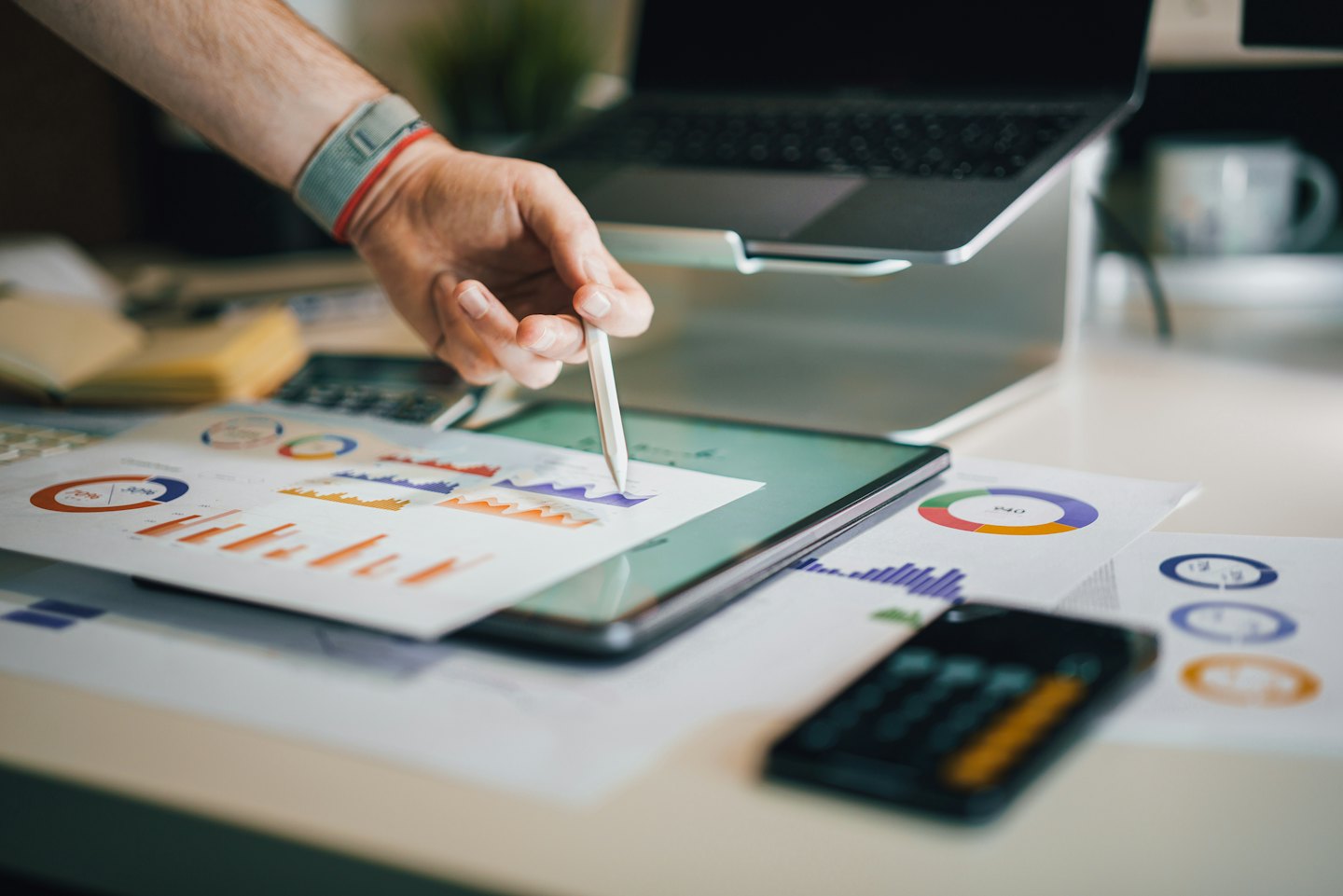 Hand with stylus points to business charts on paper over tablet.